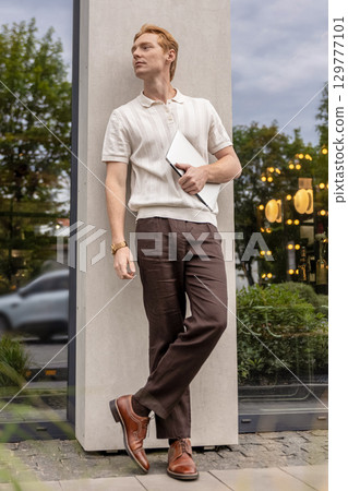 Young male with ginger hair standing quietly holding laptop near contemporary building Young male with ginger hair standing quietly holding laptop near contemporary building 129777101