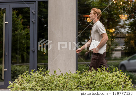 Ginger Caucasian male walking along street holding laptop near office building with greenery Ginger Caucasian male walking along street holding laptop near office building with greenery 129777123
