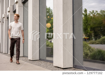 Red-haired man carrying laptop in one hand moving by the building outdoors 129777182