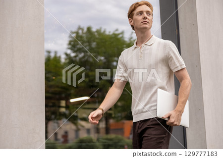 Young ginger male in his twenties walking along the street holding a laptop near modern building Young ginger male in his twenties walking along the street holding a laptop near modern building 129777183