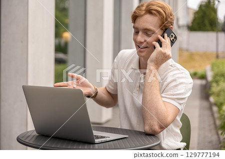 Smiling man multitasking working remotely on laptop and answering client call in open air cafe 129777194