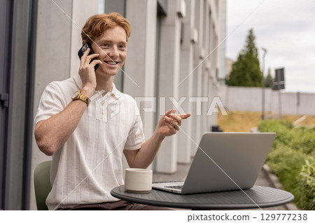 Smiling man multitasking working remotely on laptop and answering client call in open air cafe 129777238