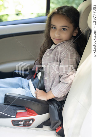 Dark-haired girl of school age sitting in a care with her seatbelt fixed 129777382