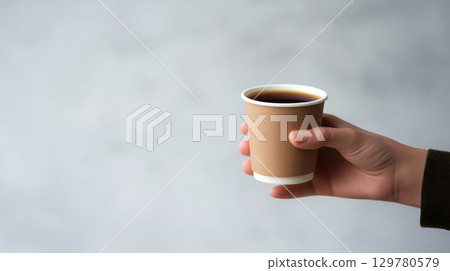 Hand holding a brown paper cup filled with black coffee against a light gray background 129780579