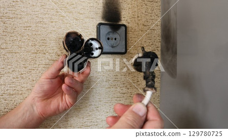 hands holding a burnt electrical plug and a scorched tee, victims of a flash and fire in a socket on the wall of a house or apartment, a dangerous situation with home electrical wiring hands holding a burnt electrical plug and a scorched tee, victims of a flash and fire in a socket on the wall of a house or apartment, a dangerous situation with home electrical wiring 129780725