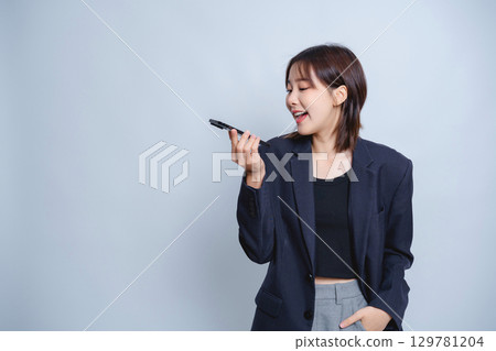 Young woman in oversized black blazer laughing while talking into voice recorder on light blue background, expressing joy and enthusiasm in conversation Young woman in oversized black blazer laughing while talking into voice recorder on light blue background, expressing joy and enthusiasm in conversation 129781204