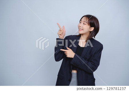 Young Asian woman in stylish blazer pointing with both hands, expressing enthusiasm and positivity, with a light blue background in a studio setting 129781237