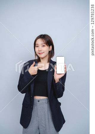 Confident Young Woman in Business Attire Pointing at Smartphone with Blank Screen, Showcasing Modern Technology and Professional Style in Minimalist Background Confident Young Woman in Business Attire Pointing at Smartphone with Blank Screen, Showcasing Modern Technology and Professional Style in Minimalist Background 129781238