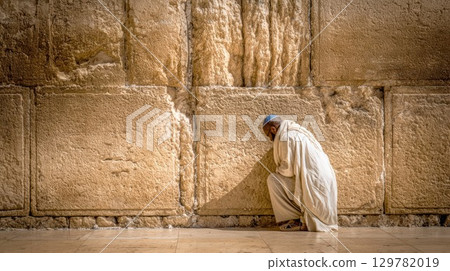 A person in traditional religious attire kneels in prayer, their head bowed against the ancient, sunlit stones of the Western Wall 129782019
