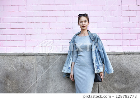 Stylish female model wearing denim jacket and grey dress. Outdoor portrait with pink wall background. Street style, fashion lifestyle and trendy urban swag look Stylish female model wearing denim jacket and grey dress. Outdoor portrait with pink wall background. Street style, fashion lifestyle and trendy urban swag look 129782778