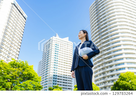 A female business person standing with a helmet in front of a city with fresh greenery A female business person standing with a helmet in front of a city with fresh greenery 129783264
