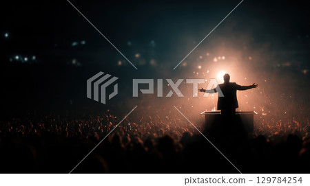 Silhouette of leader standing on elevated platform addressing large audience with dramatic backlighting in auditorium setting 129784254