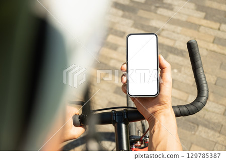 Male cyclist in helmet with mobile phone with white blank screen on bike check apps at park. Space for design. View from above. Close up. Male cyclist in helmet with mobile phone with white blank screen on bike check apps at park. Space for design. View from above. Close up. 129785387