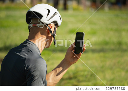 Male cyclist holding smartphone with empty blank screen on bike at park. 129785388