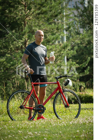Cyclist wearing gray t-shirt stand near bicycle at greenery public park. Summer outdoor activity. Vertical format Cyclist wearing gray t-shirt stand near bicycle at greenery public park. Summer outdoor activity. Vertical format 129785399