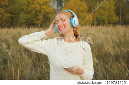 Happy young woman enjoying listening to music in headphones with phone outdoors in autumn park Happy young woman enjoying listening to music in headphones with phone outdoors in autumn park 129785825