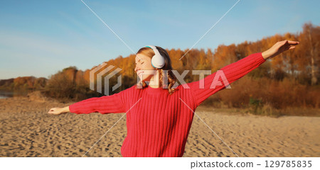 Happy young woman enjoying listening to music in headphones outdoors in autumn park 129785835