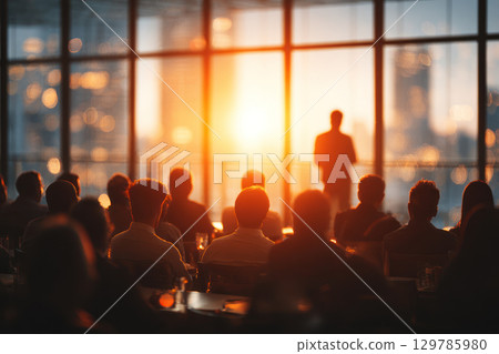 Business meeting with speaker presenting to audience in modern conference room illuminated by warm sunset light 129785980