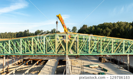Close-up front view of green timber roof trusses being installed by a mobile telescopic crane on a large concrete and brick construction site with forest in the background 129786183