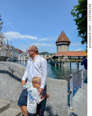A man stands beside a child near the historic Chapel Bridge in Lucerne. The child, holding a water bottle, gazes into the distance, enjoying a sunny day beside the iconic structure. 129786298