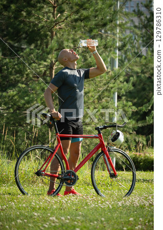 Cyclist standing with water bottle wearing gray t-shirt and relax at forest. Athlete taking a break. Outdoor activity day off. Heat summer season 129786310