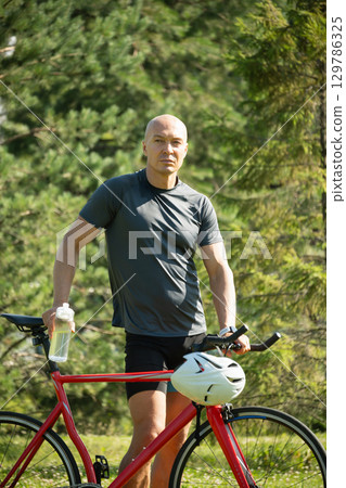 Cyclist standing with water bottle wearing gray t-shirt and relax at forest. Athlete taking a break. Outdoor activity day off. Summer season 129786325
