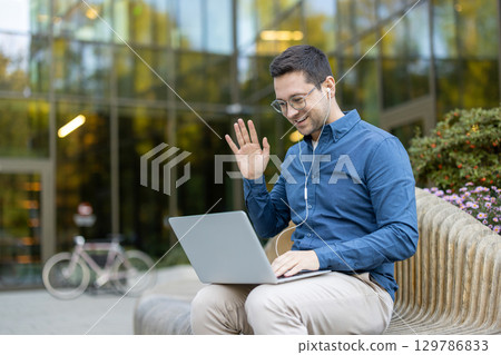 A young professional man engaging in a video call using a laptop, sitting outdoors on a wooden bench in front of a modern glass building with nearby flowers and bicycle. A young professional man engaging in a video call using a laptop, sitting outdoors on a wooden bench in front of a modern glass building with nearby flowers and bicycle. 129786833