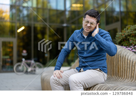 A man wearing a blue shirt and glasses sitting on a bench, holding his ear, expressing discomfort, with a blurred bicycle and modern glass building in the background. A man wearing a blue shirt and glasses sitting on a bench, holding his ear, expressing discomfort, with a blurred bicycle and modern glass building in the background. 129786848