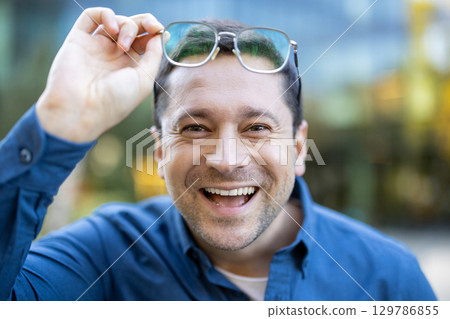 A cheerful man outdoors with a genuine smile lifting his glasses, capturing natural joy and friendliness in a casual blue shirt. The blurred background suggests an urban setting with vibrant tones. 129786855