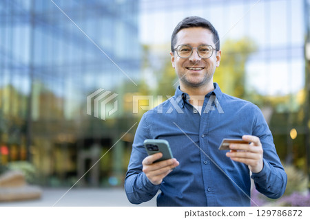 Cheerful man holding a smartphone and credit card while outdoors in a business environment. 129786872