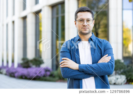 A portrait of a confident young man standing with arms crossed outdoors. Modern architecture and landscaped surroundings provide a contemporary atmosphere to the scene, reflecting professionalism A portrait of a confident young man standing with arms crossed outdoors. Modern architecture and landscaped surroundings provide a contemporary atmosphere to the scene, reflecting professionalism 129786896