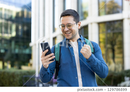 A cheerful young man wearing glasses and a backpack smiles while looking at his phone, expressing joy outdoors in a campus setting with modern architecture in the background. 129786899