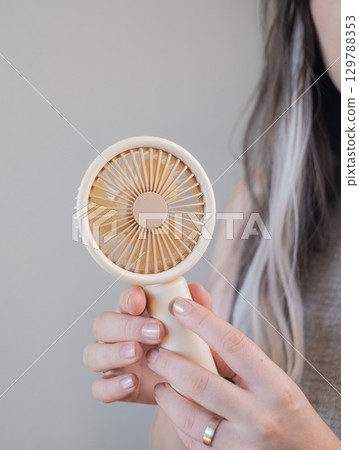 Young woman using a compact handheld fan to stay fresh and comfortable outdoors on a hot summer day. 129788353