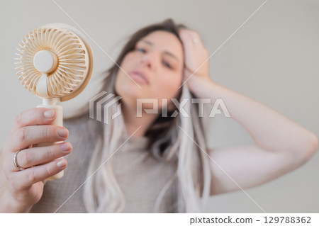 Young woman holding a mini fan to beat the heat and stay fresh outdoors on a warm sunny summer day. 129788362