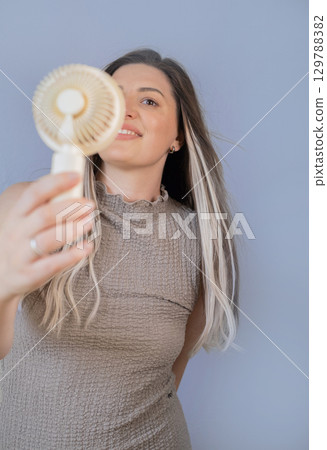 Young woman staying cool and happy with her personal portable fan on a warm sunny day outdoors. 129788382