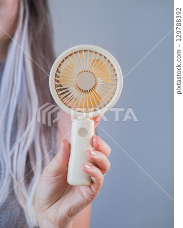 Smiling young woman staying cool with a portable fan while enjoying a warm summer day outdoors. Smiling young woman staying cool with a portable fan while enjoying a warm summer day outdoors. 129788392