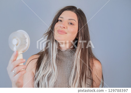 Young woman smiling while holding a portable mini fan, enjoying a cool breeze on a hot summer day. Young woman smiling while holding a portable mini fan, enjoying a cool breeze on a hot summer day. 129788397