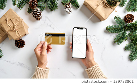Person holding gold credit card one hand and smartphone with blank screen other, surrounded by Christmas gift boxes, pine branches, and pinecones white marble surface, creating festive holiday Person holding gold credit card one hand and smartphone with blank screen other, surrounded by Christmas gift boxes, pine branches, and pinecones white marble surface, creating festive holiday 129788801