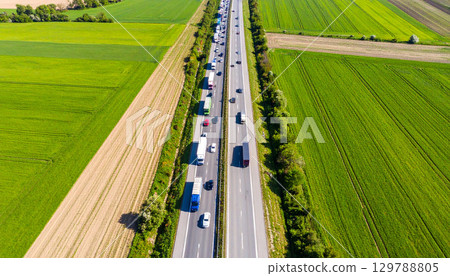 Aerial view of busy highway surrounded by lush green fields and farmland, with numerous vehicles traveling in both directions on sunny day, creating vibrant and dynamic scene Aerial view of busy highway surrounded by lush green fields and farmland, with numerous vehicles traveling in both directions on sunny day, creating vibrant and dynamic scene 129788805