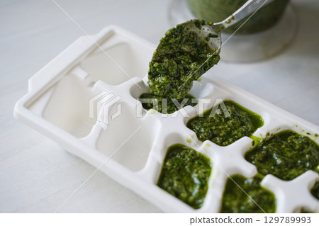 Close-up of hand using spoon to fill white ice cube tray with vibrant green pesto. Focus highlights the texture and color of sauce as it's prepared for freezing 129789993