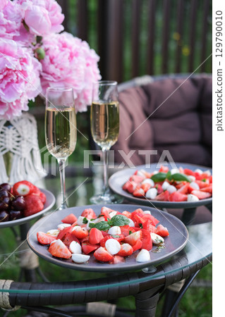 Summer garden table with two plates of strawberry, mozzarella and basil salad, champagne glasses, fresh strawberries, cherries and pink peonies. Perfect for romantic meal al fresco 129790010