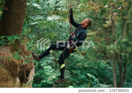 Hanging on a rope. Man is doing climbing in the forest by use of safety equipment Hanging on a rope. Man is doing climbing in the forest by use of safety equipment 129790083