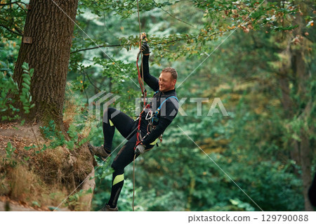 Up on the rope, hanging. Man is doing climbing in the forest by use of safety equipment Up on the rope, hanging. Man is doing climbing in the forest by use of safety equipment 129790088