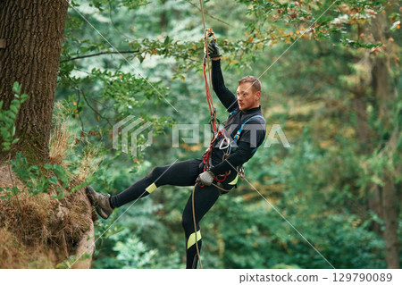 Up on the rope, hanging. Man is doing climbing in the forest by use of safety equipment Up on the rope, hanging. Man is doing climbing in the forest by use of safety equipment 129790089
