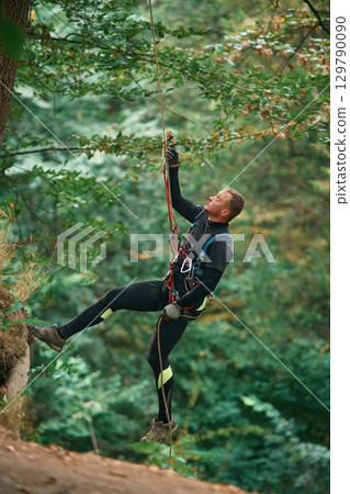 Up on the rope, hanging. Man is doing climbing in the forest by use of safety equipment Up on the rope, hanging. Man is doing climbing in the forest by use of safety equipment 129790090