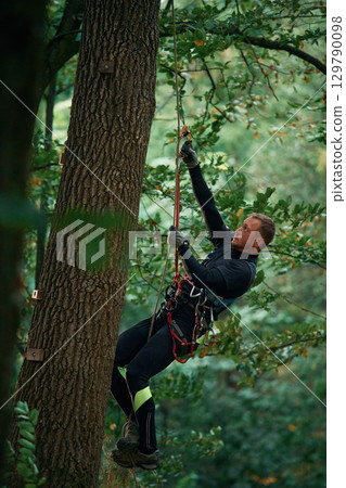 Side view, using the rope, hanging. Man is doing climbing in the forest by use of safety equipment Side view, using the rope, hanging. Man is doing climbing in the forest by use of safety equipment 129790098