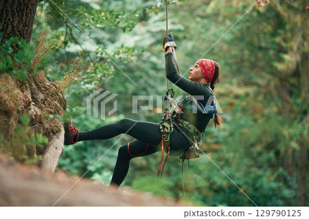 View from the side, hanging on the rope. Woman is doing climbing in the forest by the use of safety equipment View from the side, hanging on the rope. Woman is doing climbing in the forest by the use of safety equipment 129790125