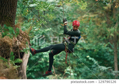 View from the side, hanging on the rope. Woman is doing climbing in the forest by the use of safety equipment View from the side, hanging on the rope. Woman is doing climbing in the forest by the use of safety equipment 129790126