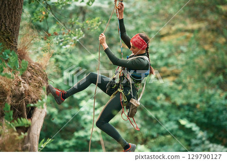 View from the side, hanging on the rope. Woman is doing climbing in the forest by the use of safety equipment 129790127