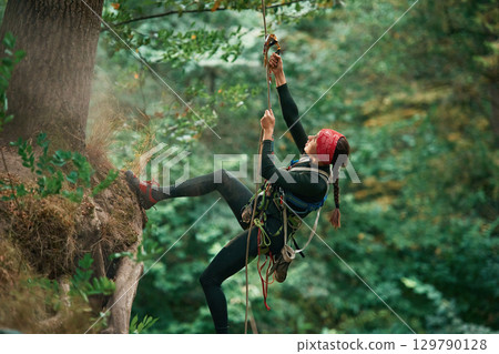 View from the side, hanging on the rope. Woman is doing climbing in the forest by the use of safety equipment View from the side, hanging on the rope. Woman is doing climbing in the forest by the use of safety equipment 129790128